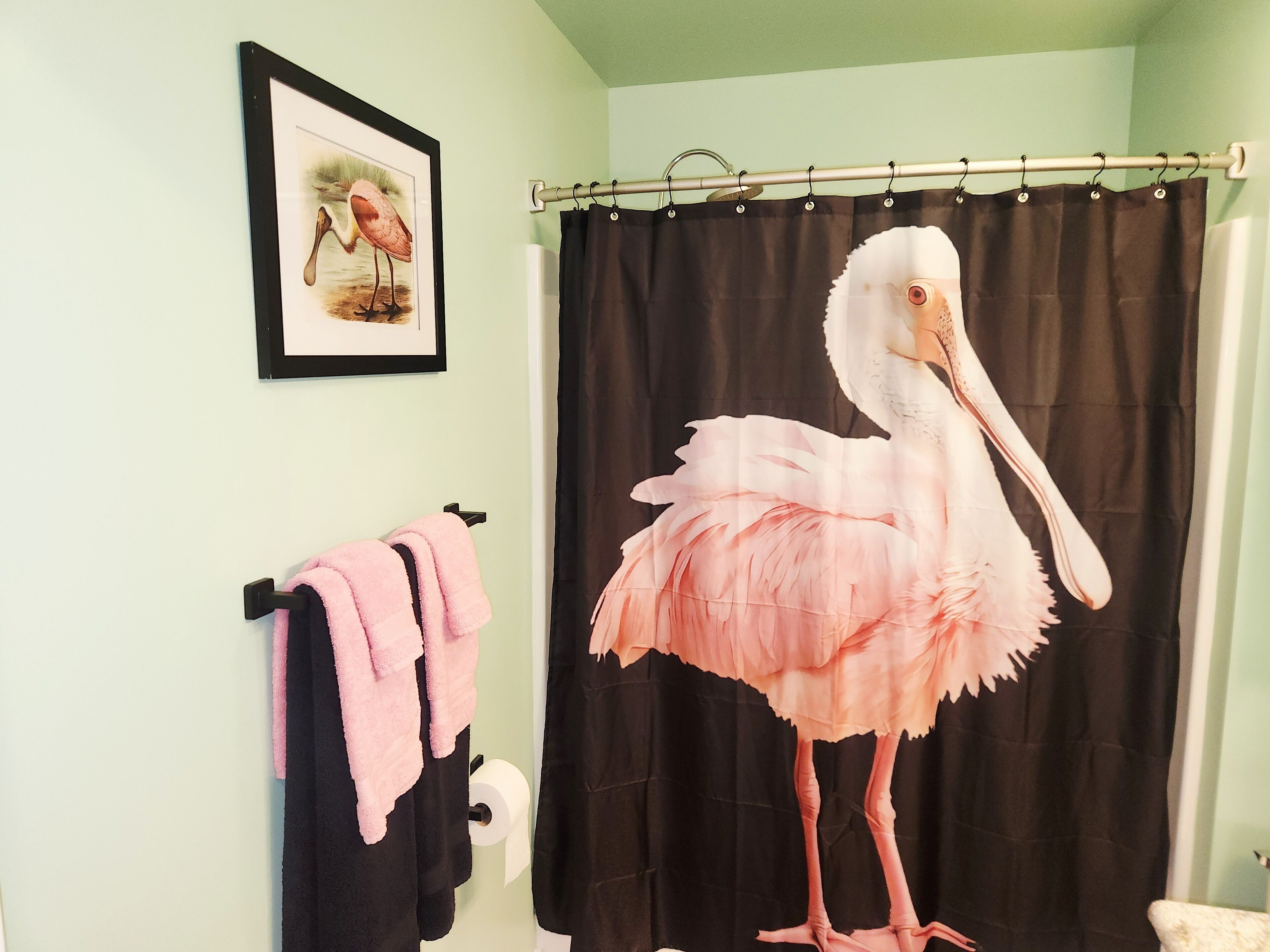 Ein Badezimmer mit einer großen, rosa Flamingo-Dekoration, einem gerahmten Bild von Flamingos und einem schwarzen Duschvorhang.