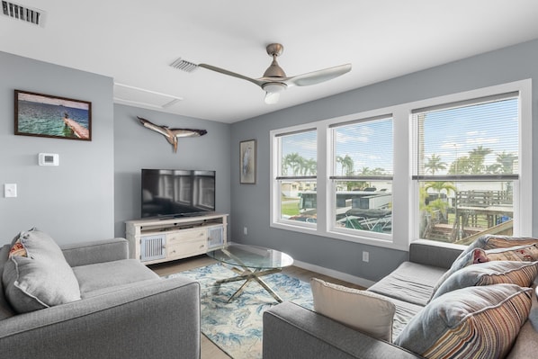 A modern living room with a flat-screen TV, a ceiling fan, and large windows with a view of the outdoors. - Amazing little fishing cabin on the water with a boat ramp in Everglades City (Everglades City)