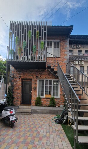 A two-story building with a brick facade, a balcony with hanging plants, a staircase, and a parked scooter.