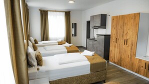Une chambre d’hôtel avec trois lits, une armoire, un coin cuisine et une grande fenêtre avec des rideaux.
