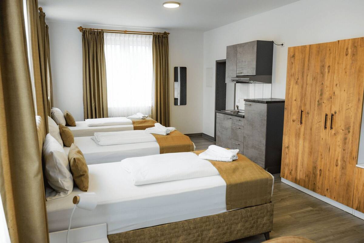 A hotel room with three beds, a wardrobe, a kitchenette, and a large window with curtains.
