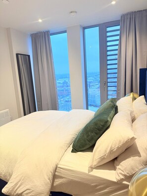 Ein Hotelzimmer mit einem großen Bett, vielen Kissen und Blick auf eine Stadtlandschaft durch die Fenster.