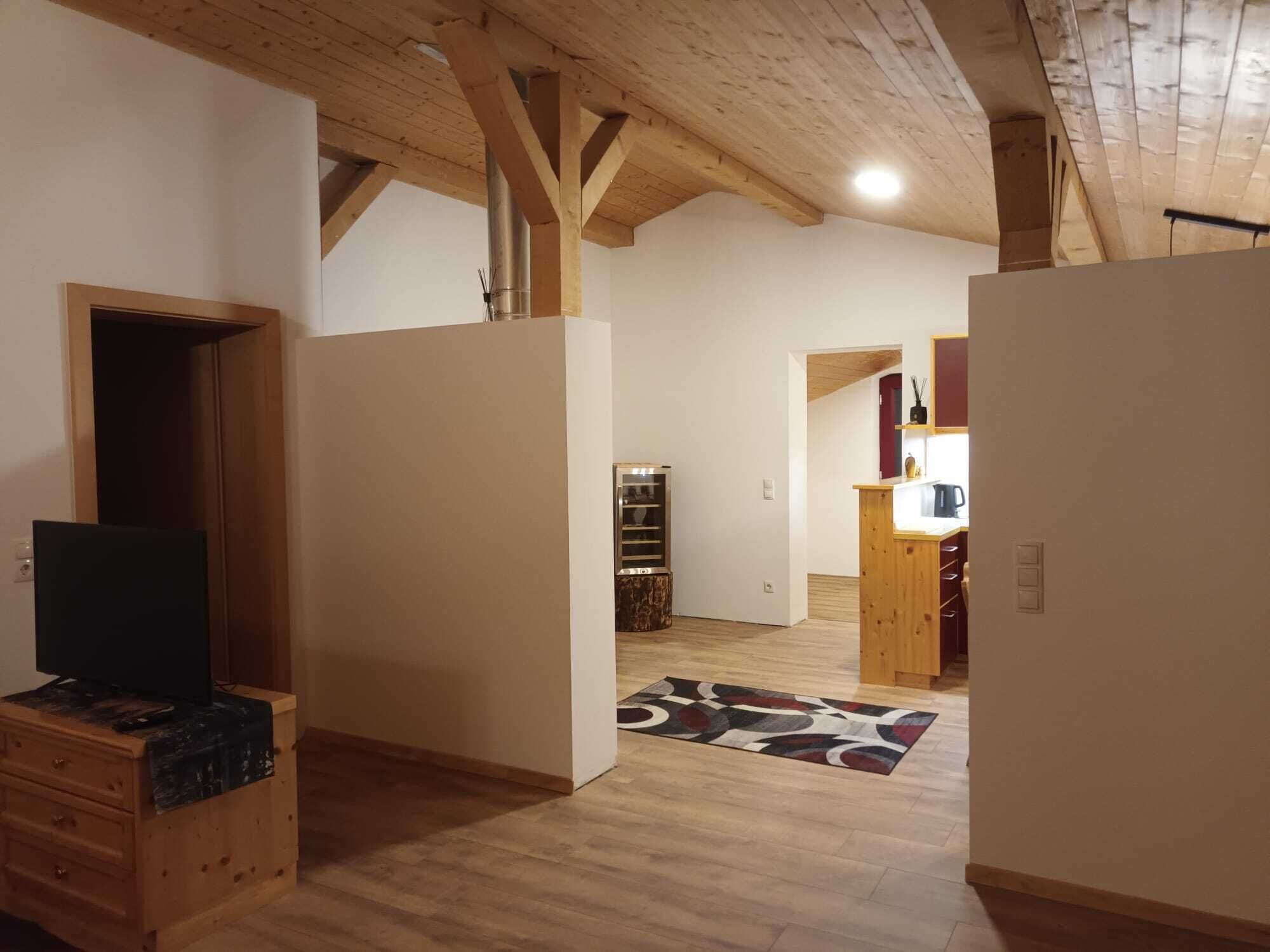 A spacious room with wooden beams, a television on a wooden dresser, and a kitchen area with a wooden counter and shelves.