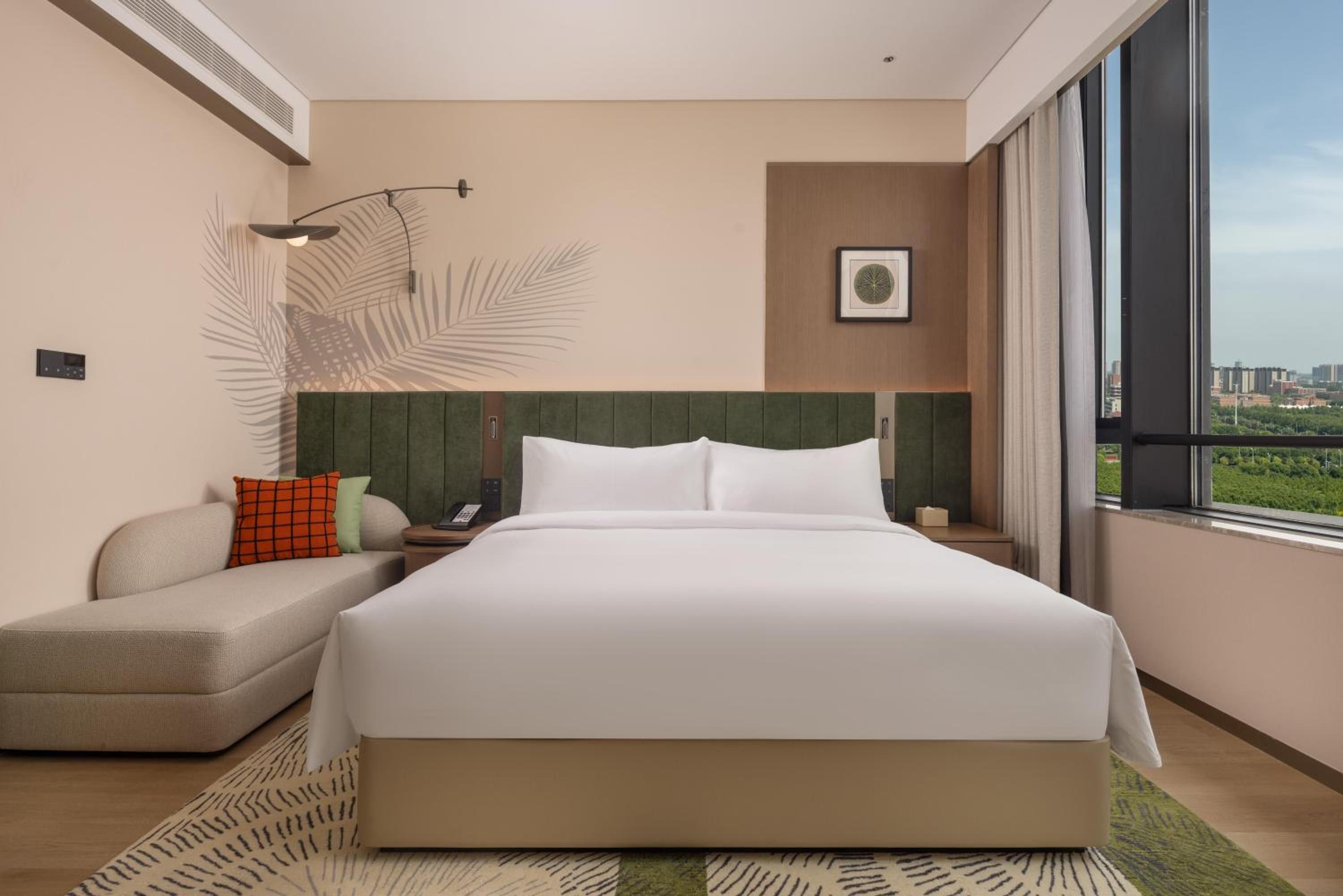 A modern hotel room with a large bed, a seating area, a wall-mounted phone, and a window with a city view.