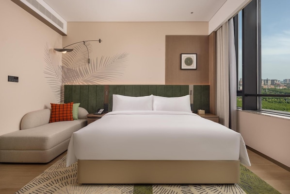 A modern hotel room with a large bed, a seating area, a wall-mounted phone, and a window with a city view.