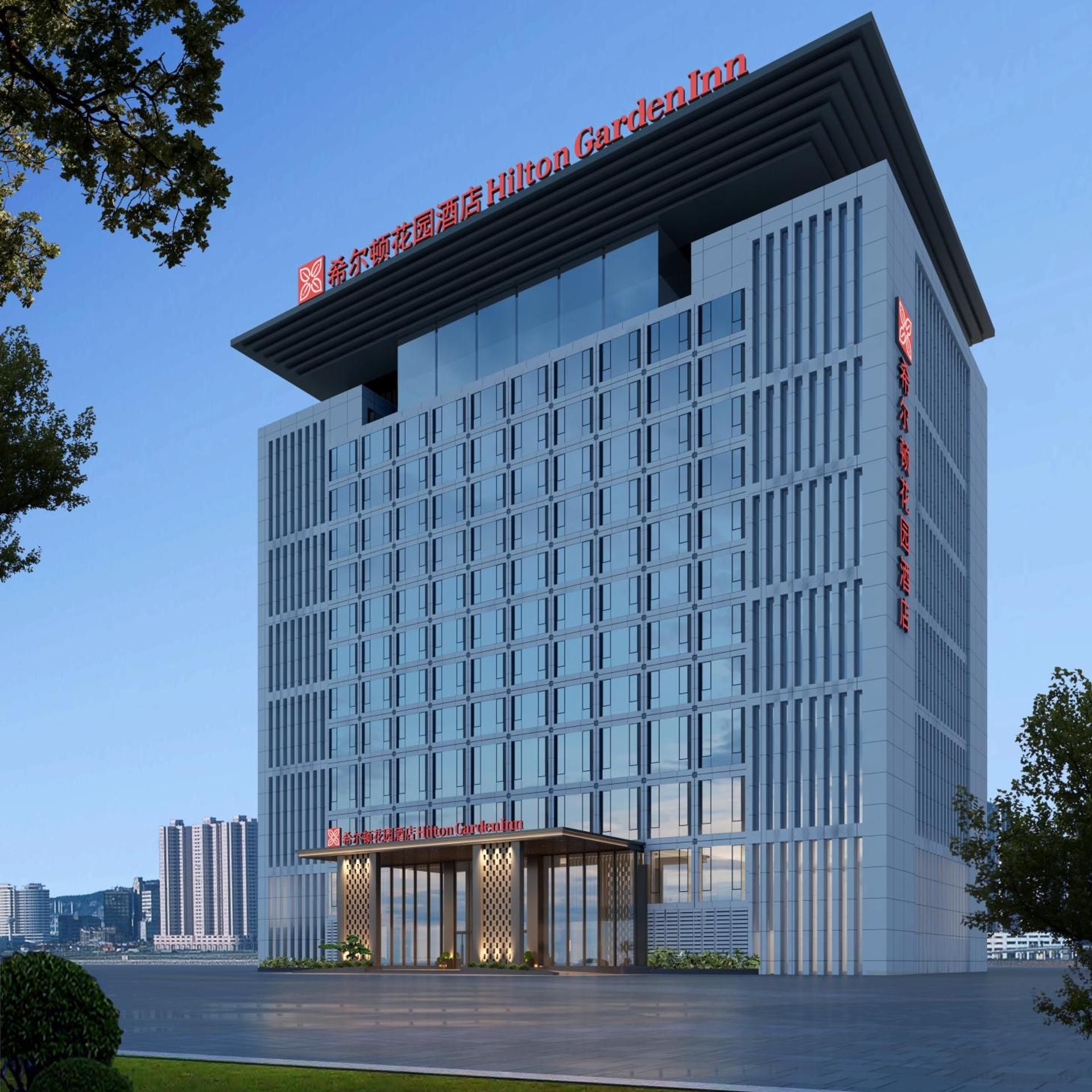 A modern hotel building with a clear glass facade, a prominent entrance with a covered area, and signage indicating it is a Hilton Garden Inn.