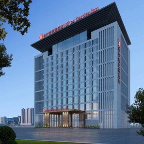 A modern hotel building with a clear glass facade, a prominent entrance with a covered area, and signage indicating it is a Hilton Garden Inn.