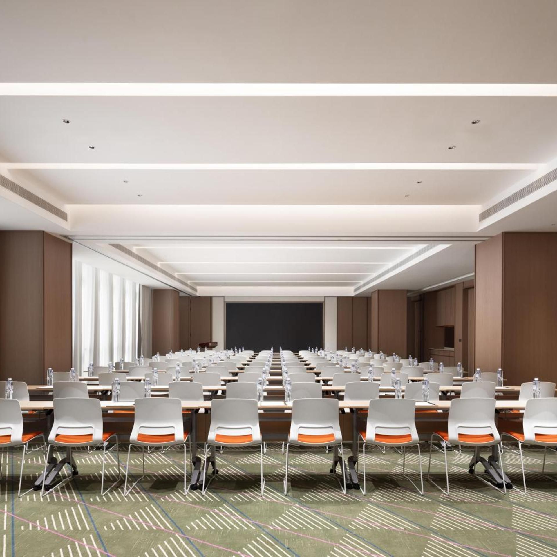 A conference room with rows of tables and chairs, a projector screen, and a carpeted floor.