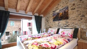 Ein Schlafzimmer mit Steinwand, Holzbalkendecke, einem Bett mit floraler Bettwäsche und einem Gemälde darüber.