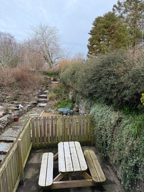 Puinen piknikpöytä, jossa on penkit, kivimuurin ja tiheän pensaikon ympäröimänä.