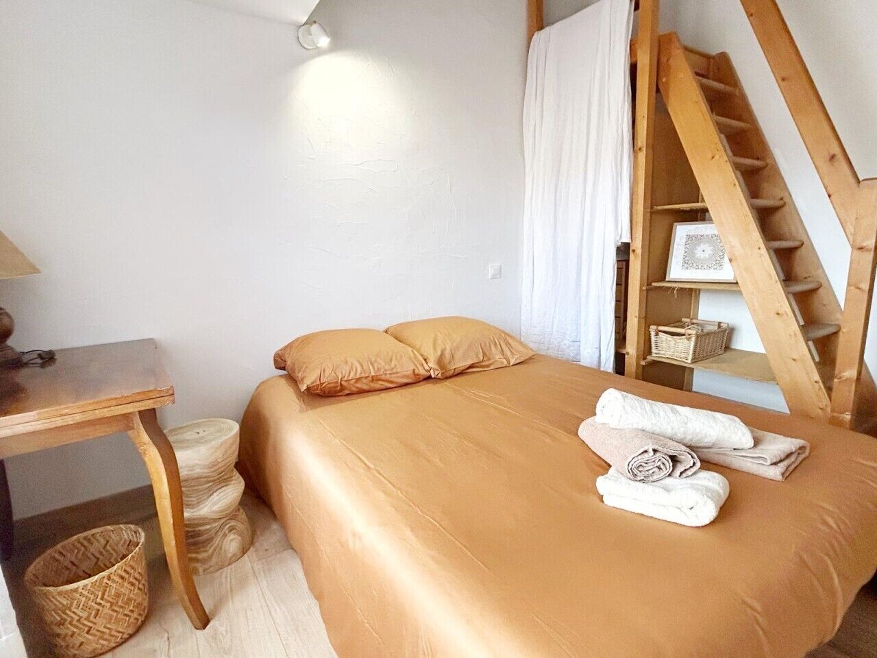 Ein Schlafzimmer mit einem Bett, Kissen, einem Holztisch, einem Stuhl, einem kleinen Korb und einer Holztreppe.