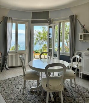 Ein Essbereich mit einem runden Tisch, vier Stühlen, großen Fenstern mit Vorhängen, einem Heizkörper und Blick aufs Meer.