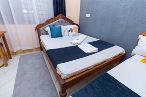 A double bed with a wooden frame, a dark blue bedspread, and white bedding. There is a wooden nightstand to the left and a small table with a chair to the right. - Dondoo's Paradise Kilifi (Kilifi)