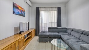 En moderne stue med en hjørnesofa, et glasbord, et fladskærms-tv på en trækommode og et stort vindue med lette gardiner.