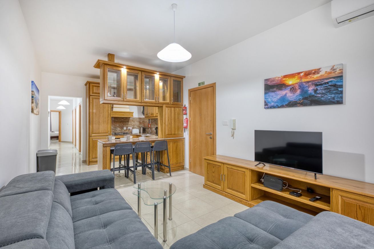 Una sala de estar moderna con un sofá gris, una mesa de centro de vidrio, un televisor sobre un mueble de madera y una cocina al fondo.