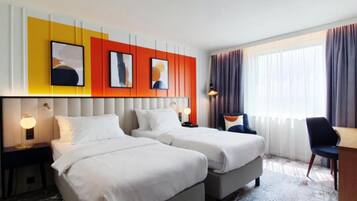 Ein Hotelzimmer mit zwei Betten, einem Schreibtisch und einem Stuhl. Zwei Nachttische mit Lampen stehen bereit. Das Zimmer verfügt über ein großes Fenster mit Vorhängen.