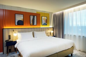 A hotel room with a large bed, two bedside tables with lamps, a headboard with artwork, and a window with curtains. - Voco Dusseldorf Seestern An Ihg Hotel (Düsseldorf)