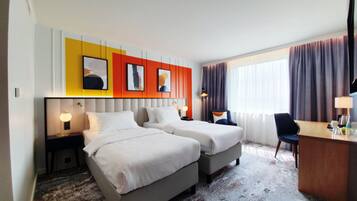 Een hotelkamer met twee bedden, een bureau en een stoel. Er zijn wandlampen en een groot raam met gordijnen.
