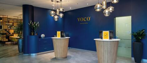 Zona de recepción con dos mostradores de madera, una pared azul con el nombre del hotel y lámparas colgantes esféricas.