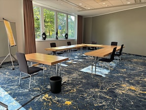 A conference room with a large table, chairs, a projector stand, and a view of trees through the windows. - Voco Dusseldorf Seestern An Ihg Hotel (Düsseldorf)
