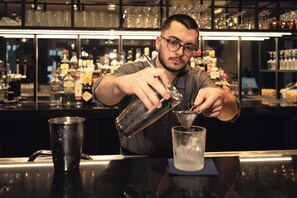 Un bartender preparando una bebida en la barra, con estantes llenos de botellas y vasos al fondo.