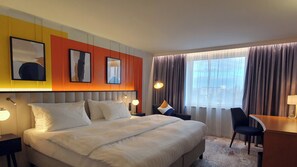A hotel room with a large bed, bedside tables, a desk, and a chair. There are two framed pictures on the wall, a window with curtains, and a view of buildings outside. - Voco Dusseldorf Seestern An Ihg Hotel (Düsseldorf)