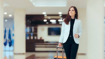 Eine Person in einem weißen Blazer und schwarzer Hose hält einen Koffer in einer Hotelhalle mit modernem Interieur und einem flachbildschirmfernseher an der Wand.