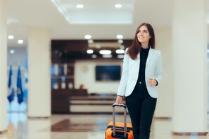 Una persona con un blazer blanco y pantalones negros sostiene una maleta en el vestíbulo de un hotel con una decoración moderna y un televisor de pantalla plana en la pared.