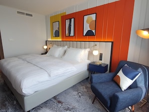 A modern hotel room with a large bed, a nightstand with a lamp, a blue armchair, and abstract art on the wall. - Voco Dusseldorf Seestern An Ihg Hotel (Düsseldorf)