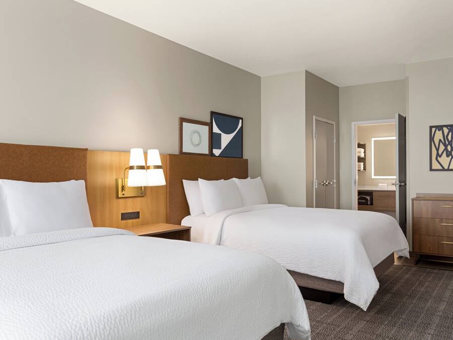A hotel room with two beds, a nightstand with a lamp, a wooden headboard, a dresser, and a door leading to a bathroom.