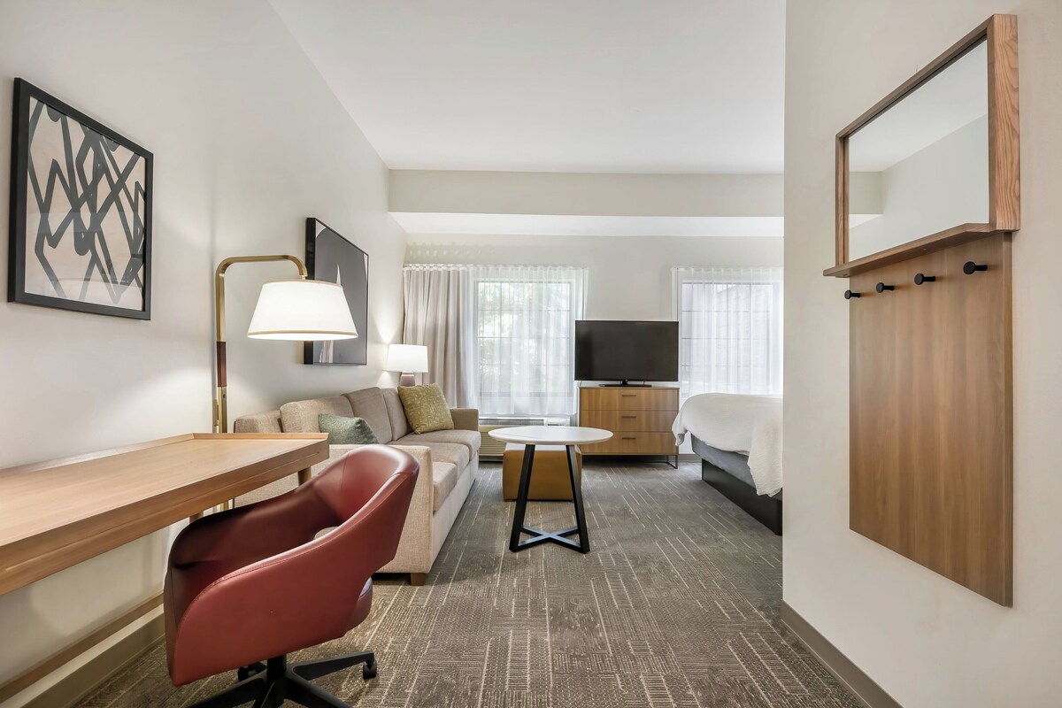 Une chambre d’hôtel avec un bureau, une chaise rouge, un canapé, une télévision, un lit et un miroir.