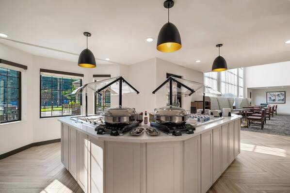 A modern kitchen with a central island, overhead pendant lighting, and a view of the outdoors through large windows. - Staybridge Suites Detroit North - Royal Oak by IHG (Troy)
