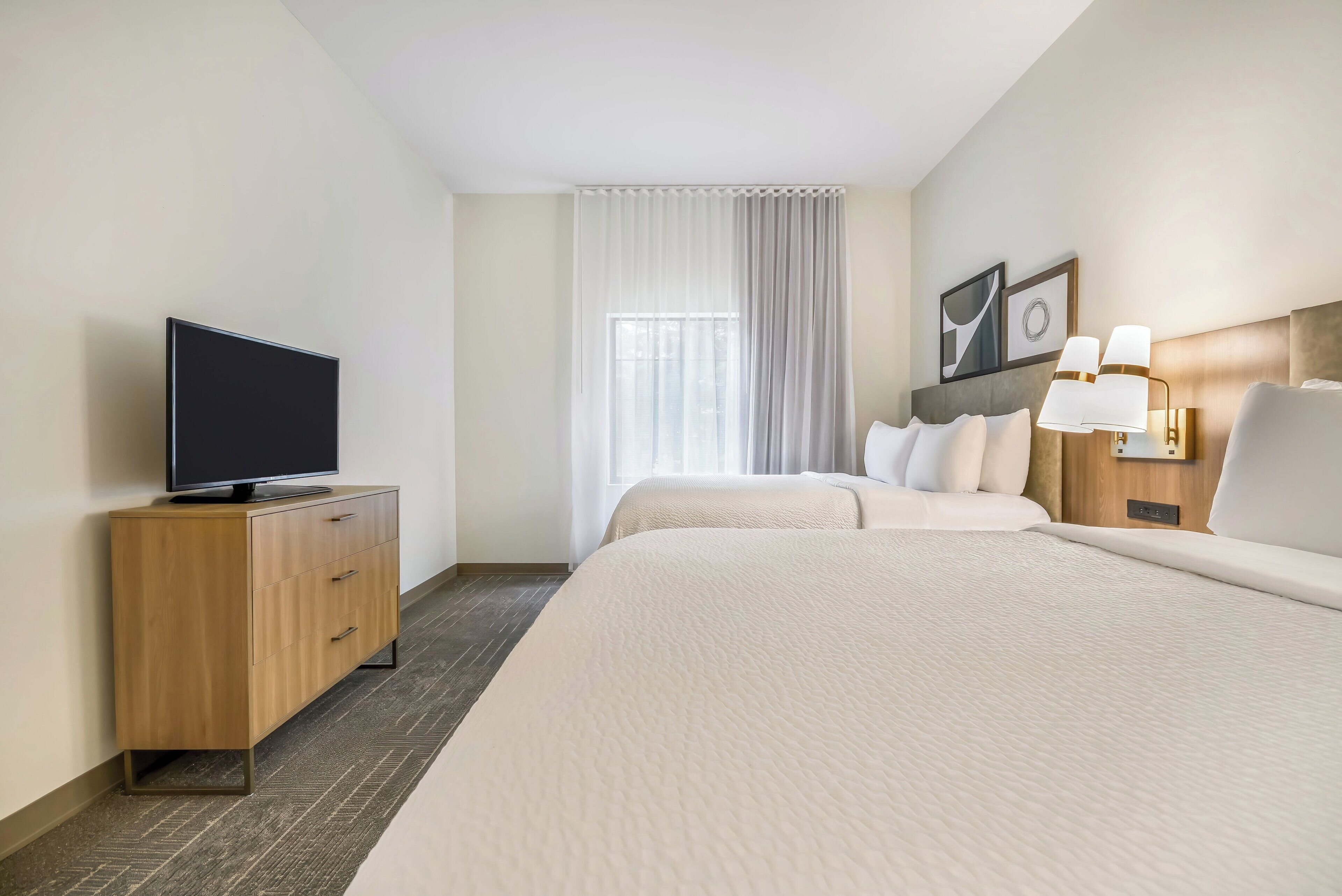 Une chambre d’hôtel avec un téléviseur à écran plat posé sur un meuble en bois, un lit avec une literie blanche, une table de chevet avec une lampe et une fenêtre ornée de rideaux légers.