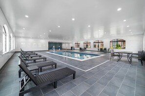 Ein modernes Hallenbad mit Liegestühlen und Tischen, umgeben von großen Fenstern und einer hohen Decke.