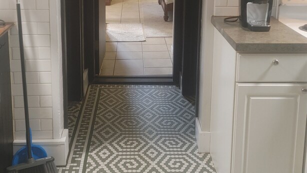 A narrow hallway with patterned floor tiles, leading to a bedroom with a bed and dresser. A coffee maker is visible on the counter.