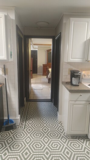 A narrow hallway with patterned floor tiles, leading to a bedroom with a bed and dresser. A coffee maker is visible on the counter.