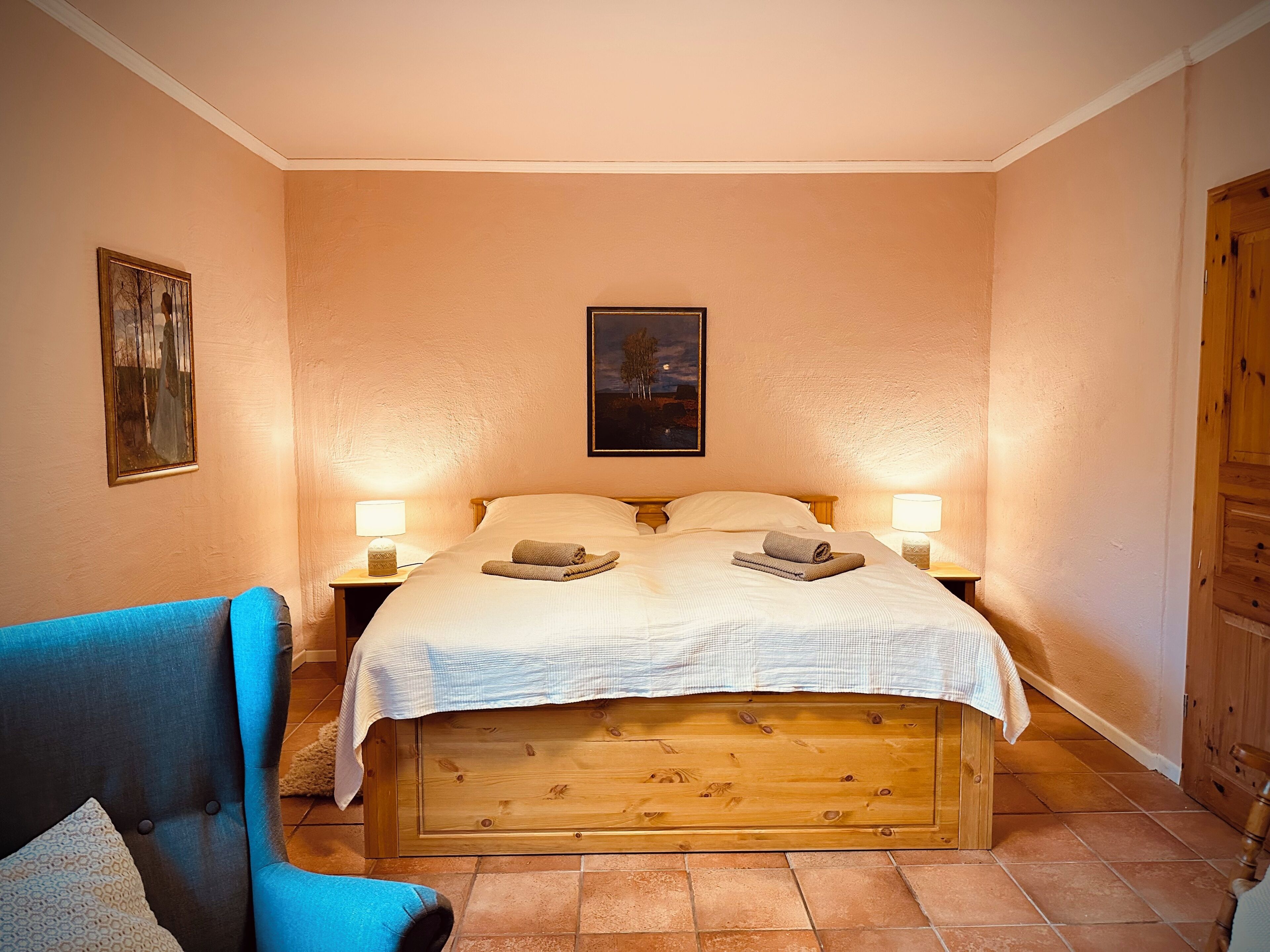 Une chambre à coucher avec un lit en bois, deux lampes de chevet, un fauteuil bleu et un tableau accroché au mur.