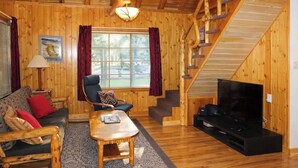 Interior de una cabaña de madera con sofá, sillón, mesa de centro, televisor y una escalera.