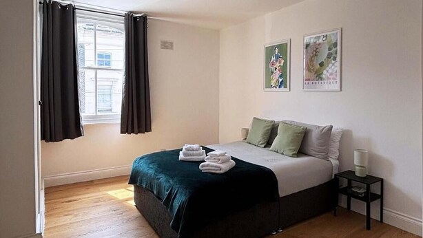 Un dormitorio con cama, mesita de noche, ventana con cortinas y dos cuadros en la pared.