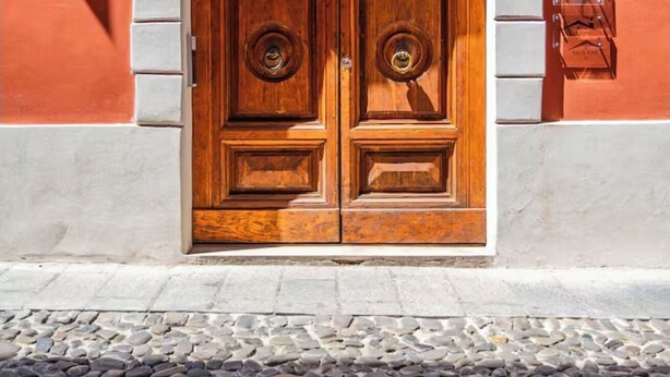 Uma porta dupla de madeira com arco de pedra, embutida em um prédio vermelho, com uma rua de paralelepípedos à frente.