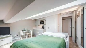 Quarto compacto com cama, escrivaninha e pequeno espaço de cozinha.