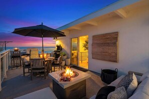 A modern outdoor patio with a fire feature, seating, and a view of the ocean at dusk. - Oceanview Luxe Home | Steps to the Strand | Bikes (Manhattan Beach)