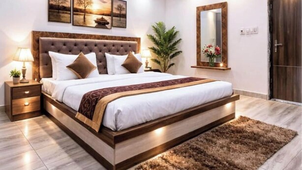 A bedroom with a large bed, a nightstand, a lamp, a mirror, and a plant. - Hotel Uday Raj (Agra)