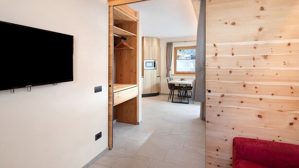 Een modern interieur met een flatscreen televisie aan de muur, een houten garderobe, een eethoek met een tafel en stoelen, en een rode bank.