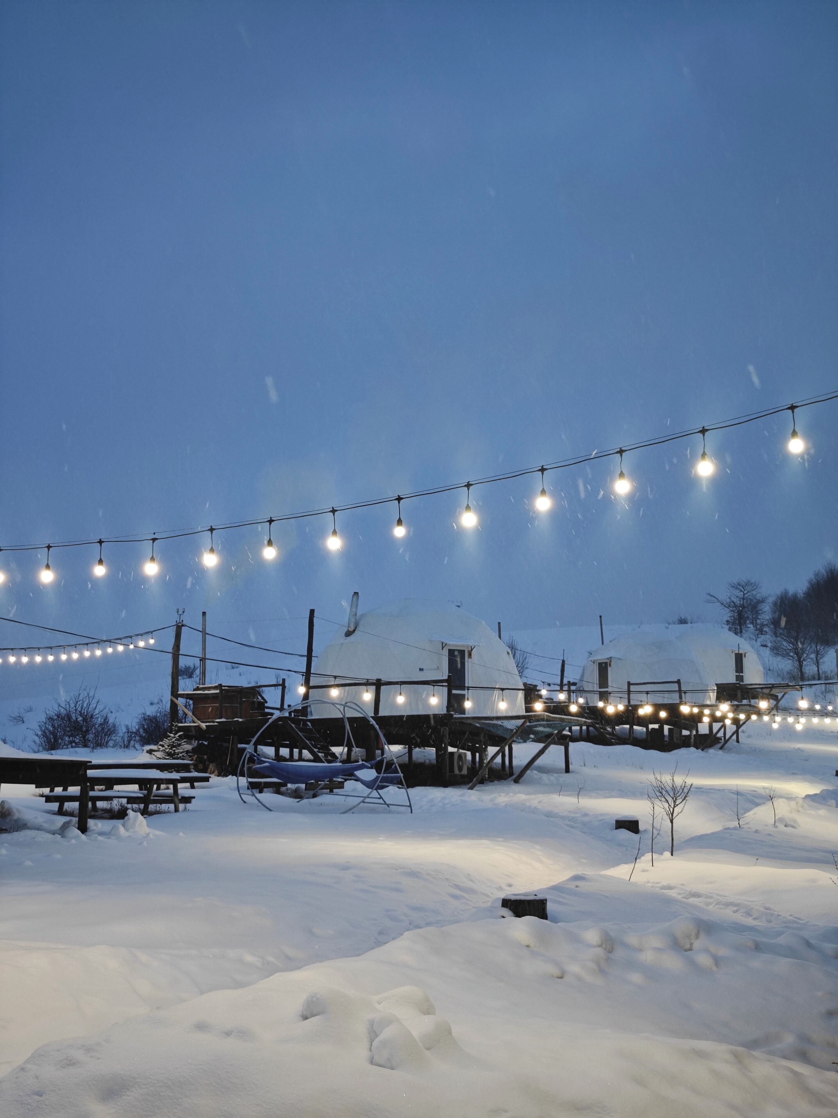 A snow-covered outdoor area with string lights, wooden structures, and a dome-shaped building.