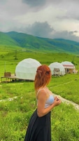 Una mujer de pie en un campo con tres estructuras en forma de cúpula al fondo.