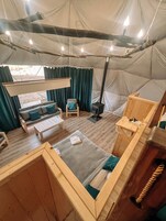 Interior de una tienda de campaña con sofá, mesa, zona de dormir y estufa de leña.