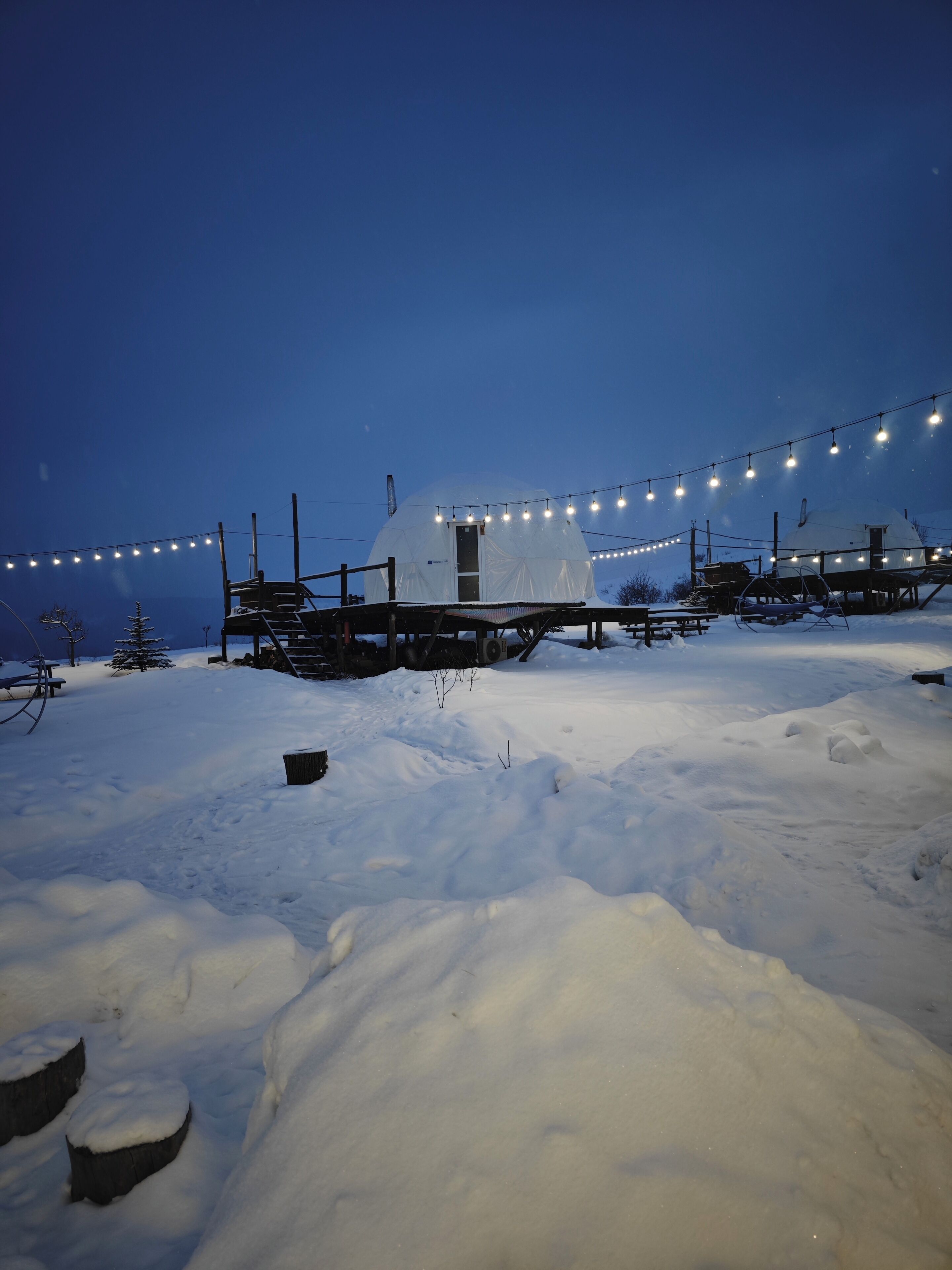 A snow-covered landscape with a white dome structure, possibly a yurt, and a string of lights.