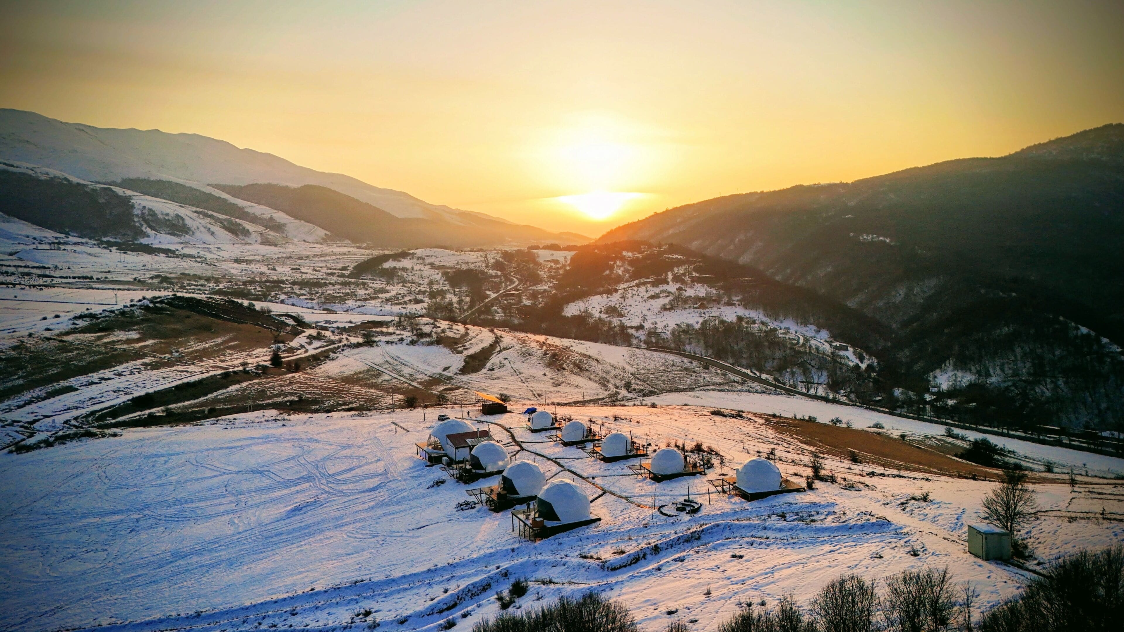 A snow-covered landscape with spherical structures, likely a resort or retreat, set against a backdrop of mountains and a setting sun.