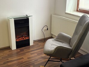 A room with a wood-burning stove, a grey upholstered chair, and a radiator. - Vacation apartment Reitzenhain (Marienberg)
